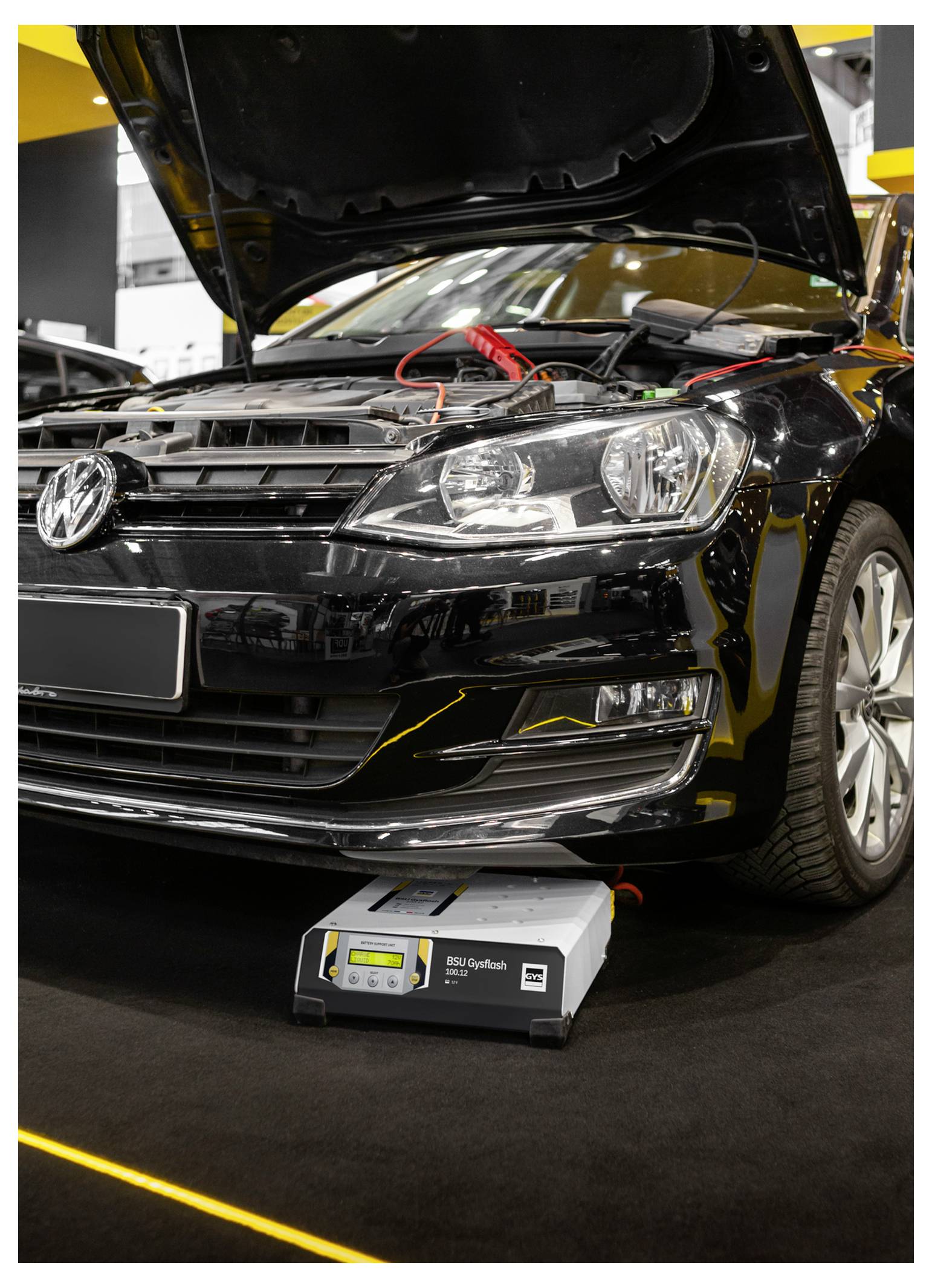 Černý vůz Volkswagen s otevřenou kapotou a připojenou nabíječkou baterie v dílně nebo na automobilové výstavě.