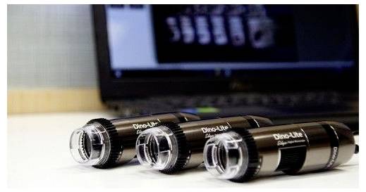 Tři ruční mikroskopy Dino-Lite jsou položeny na stole s rozmazaným laptopem v pozadí, který zobrazuje nejasný obraz.