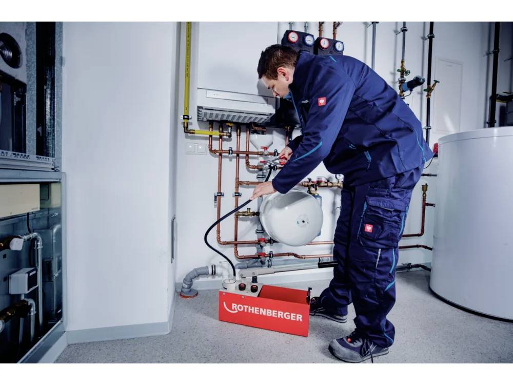 Technik v modré uniformě používá vybavení pro práci na potrubí v technické místnosti. Na podlaze leží červená montážní brašna Rothenberger.