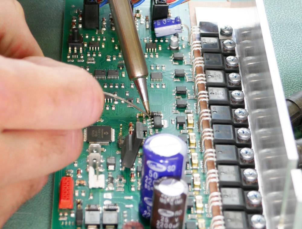 Osoba pájí komponenty na zelenou desku plošných spojů osazenou různými elektronickými součástkami.