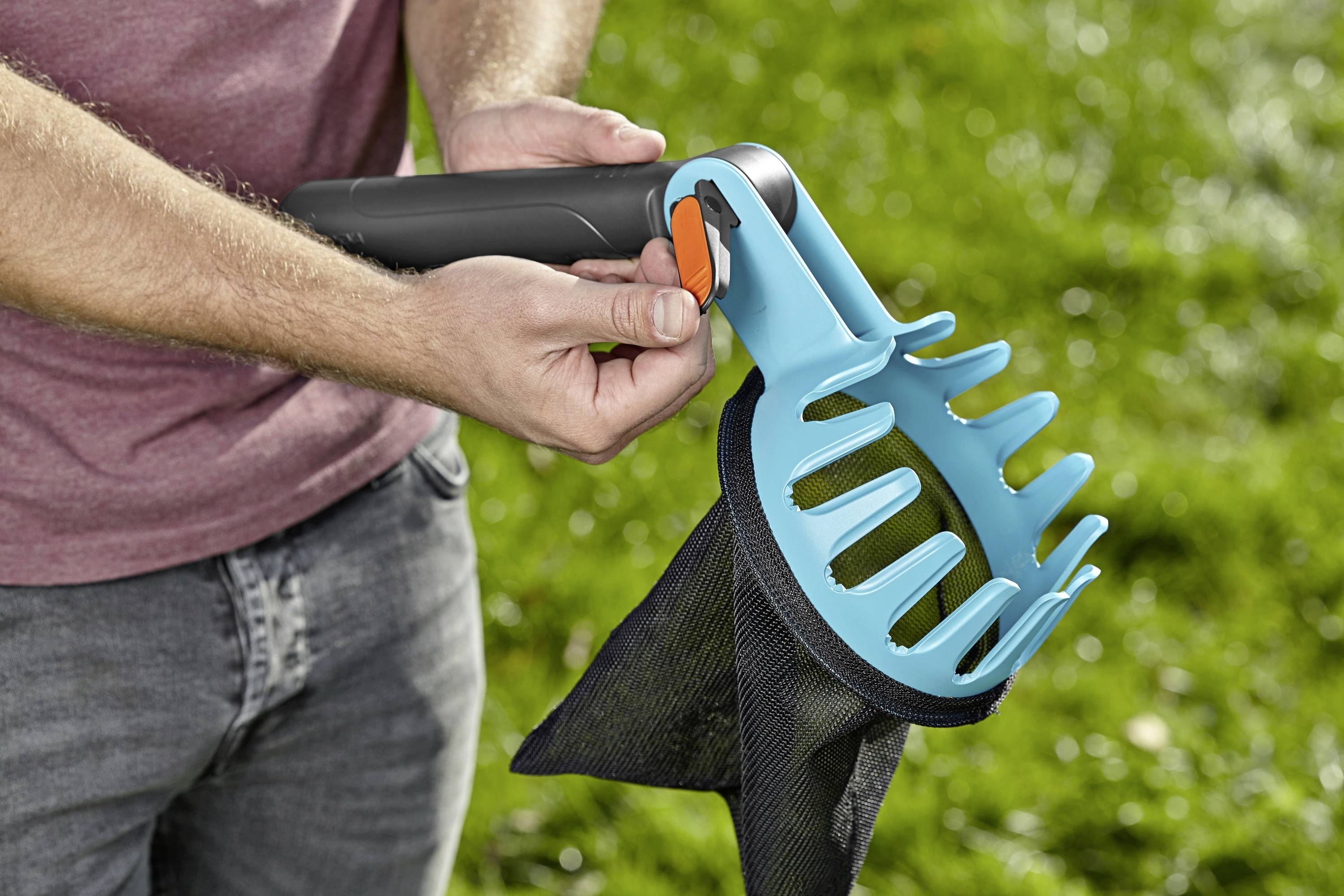 Osoba drží modrý sběrač ovoce s pytlíkem nad zelenou travnatou plochou, připravená sklízet ovoce.