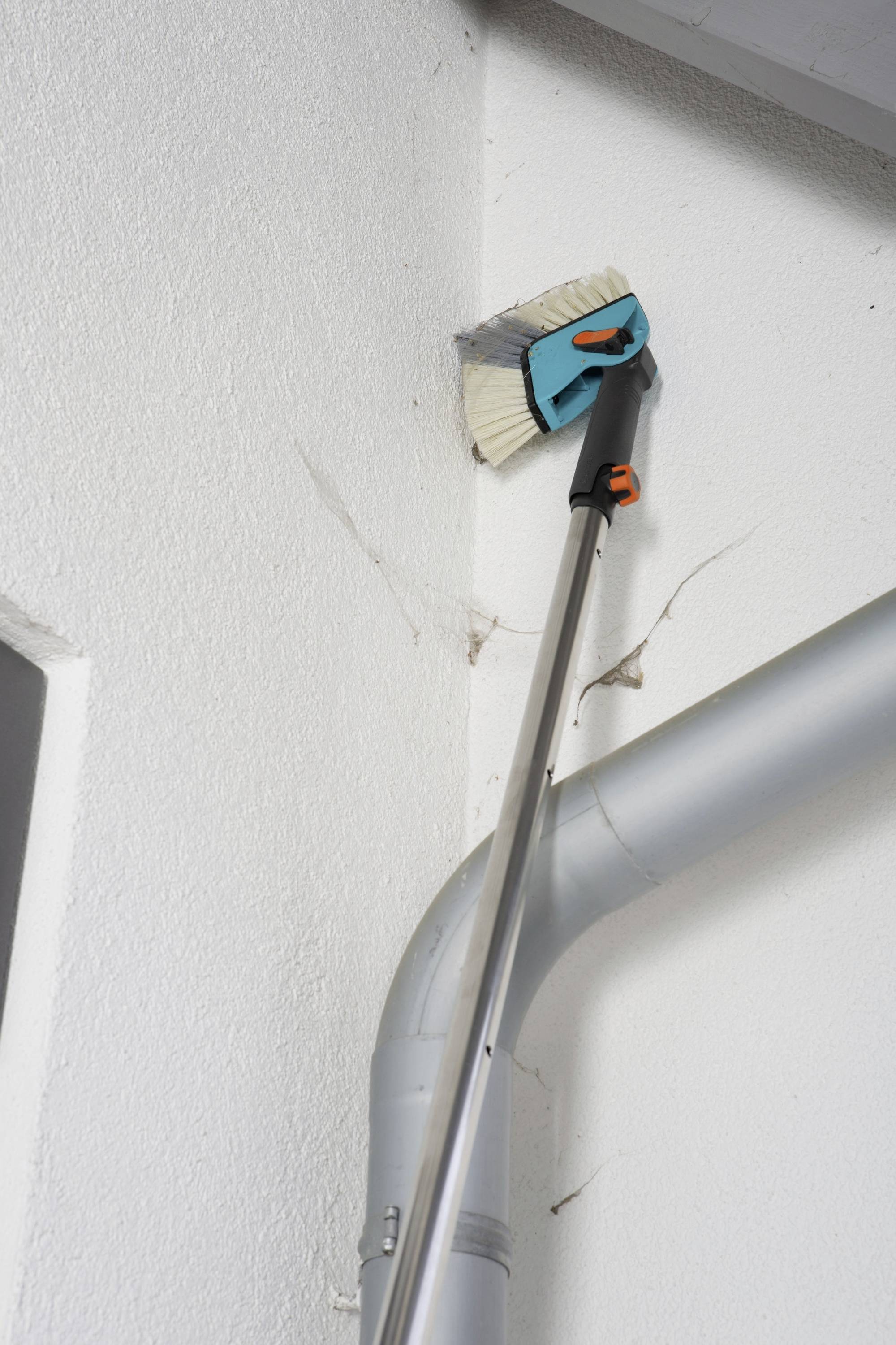 Osoba čistí pavučiny na stropě místnosti teleskopickým smetákem vedle šedého potrubí.