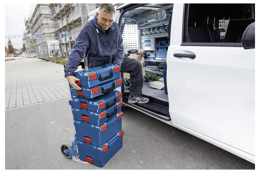 Osoba skládá věž modrých a červených nářaďových boxů na vozík vedle otevřených dveří dodávky, což naznačuje pracovní situaci nebo organizaci nástrojů.