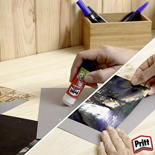 Dvě ruce používající lepicí tyčinku Pritt k připevnění krajinných fotografií na šedý papír na dřevěném stole.