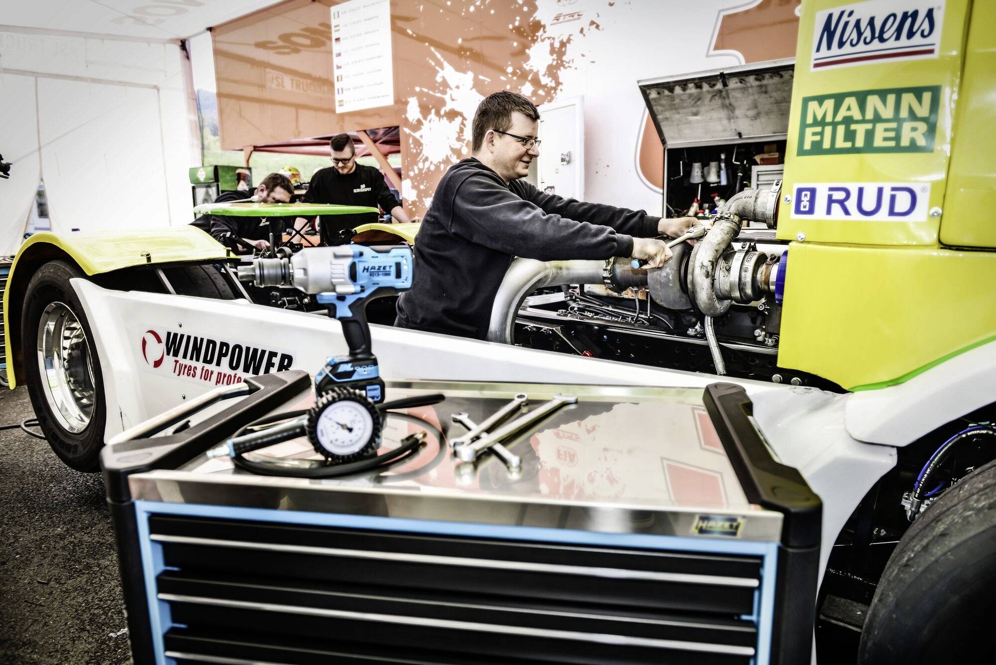 Muž pracuje na motoru závodního vozu v dílně, obklopen nástroji a náhradními díly.