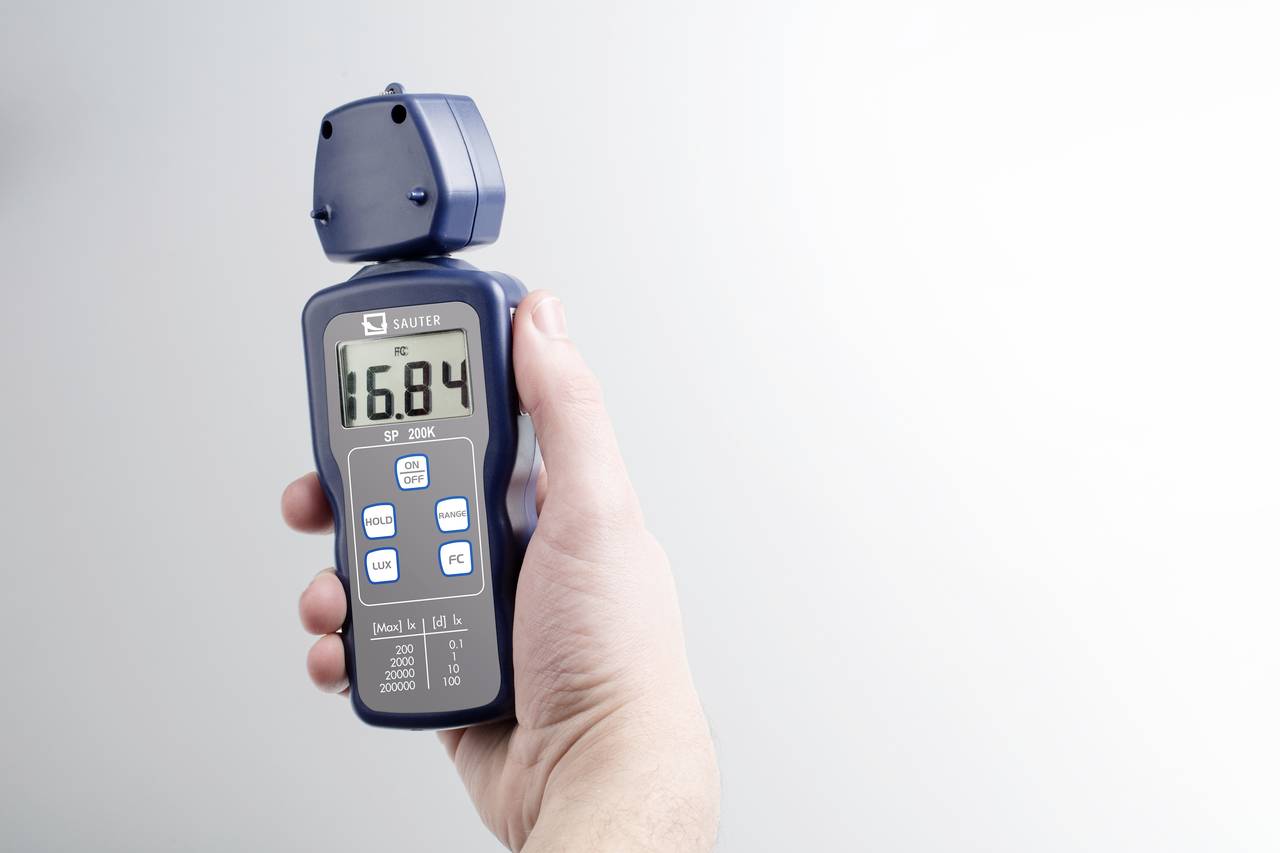 Jedna ruka drží digitální anemometr, který ukazuje hodnotu 16,84. Přístroj měří rychlost větru a teplotu.