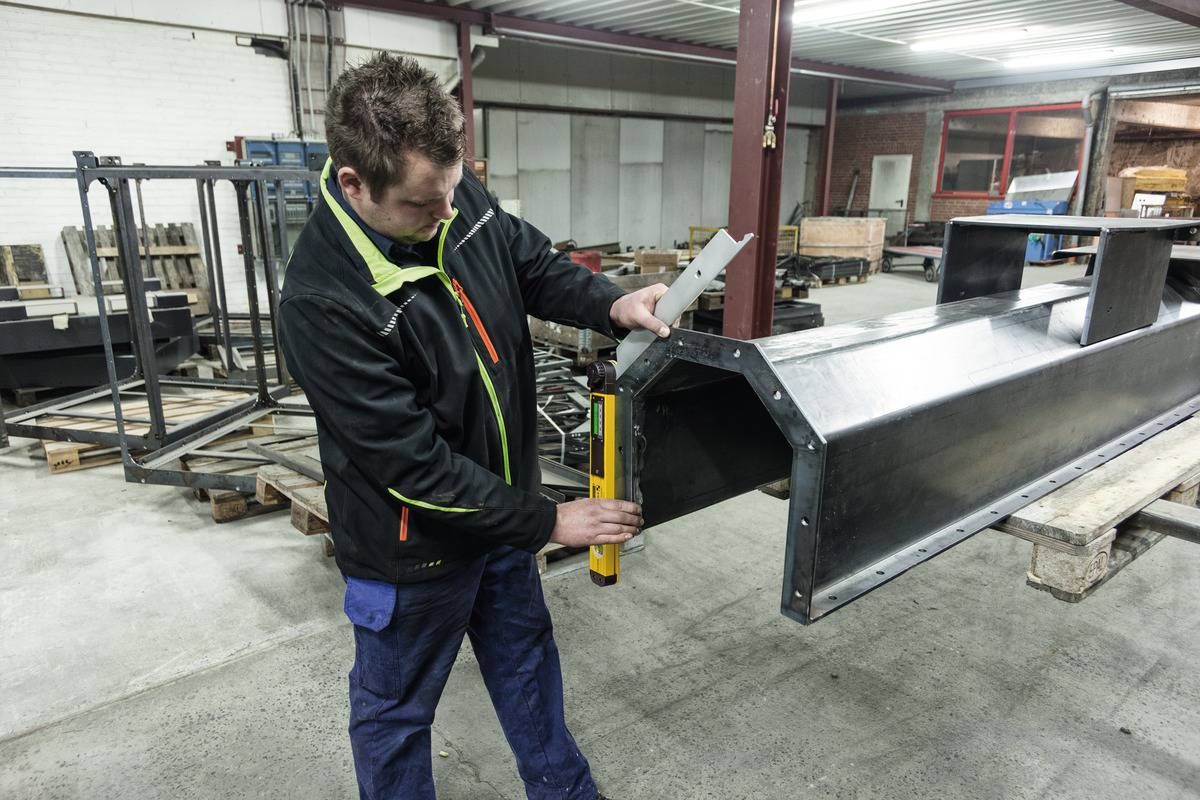 Muž měří kovovou strukturu laserovým měřícím přístrojem v průmyslové hale. V pozadí jsou viditelné stroje a materiály.