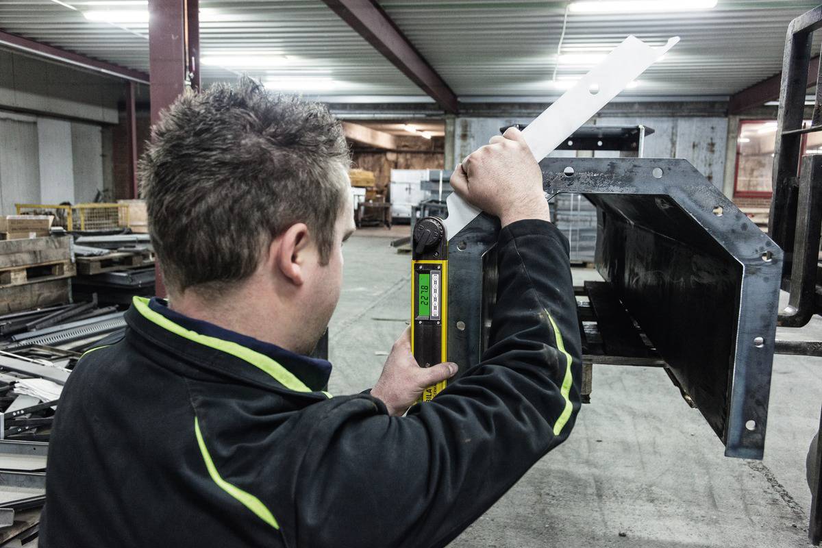 Osoba měří délku bílého kovového dílu digitálním měřicím přístrojem v dílně. V pozadí jsou regály skladu.