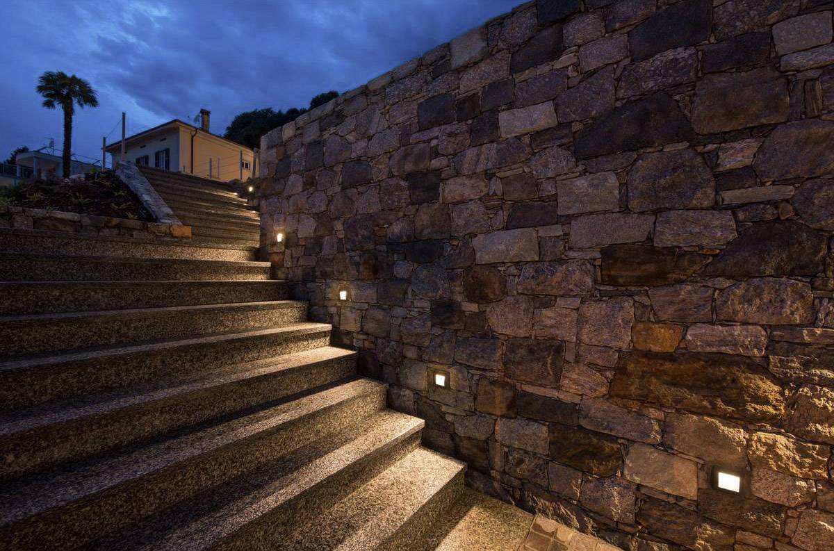 Schodiště za soumraku s osvětlenými schody a kamennou zdí vpravo. Nahoře je vidět dům s palmou nalevo.