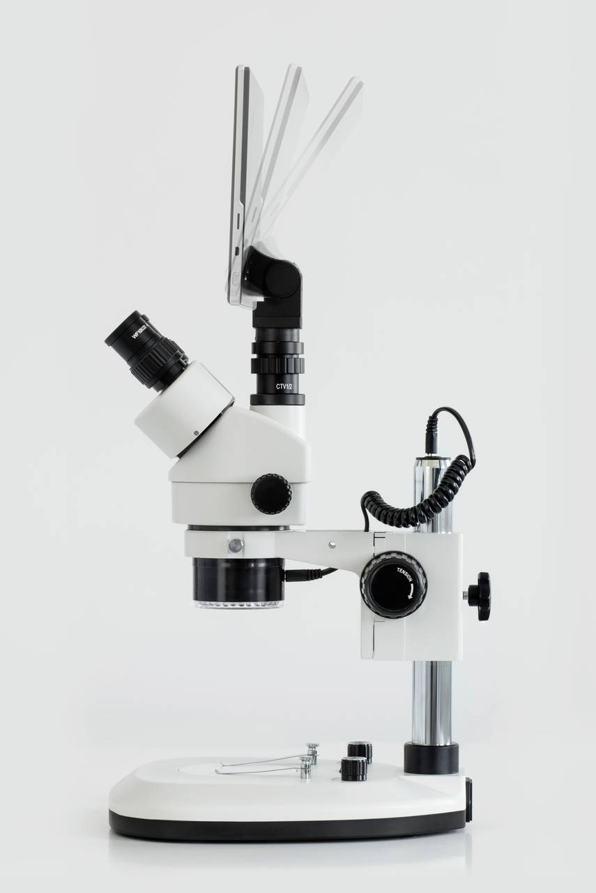 Bílý stereomikroskop s černým okulárem a osvětlením, umístěný na stabilním stojanu před neutrálním pozadím.