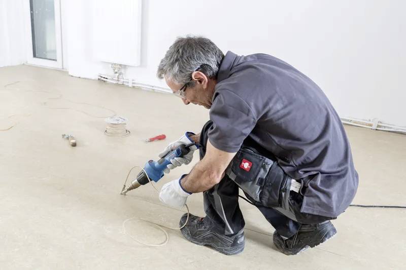 Osoba klečící na podlaze používá elektrickou vrtačku k práci na povrchu. Kolem jsou roztroušeny nástroje a dráty, což naznačuje úkol opravy.