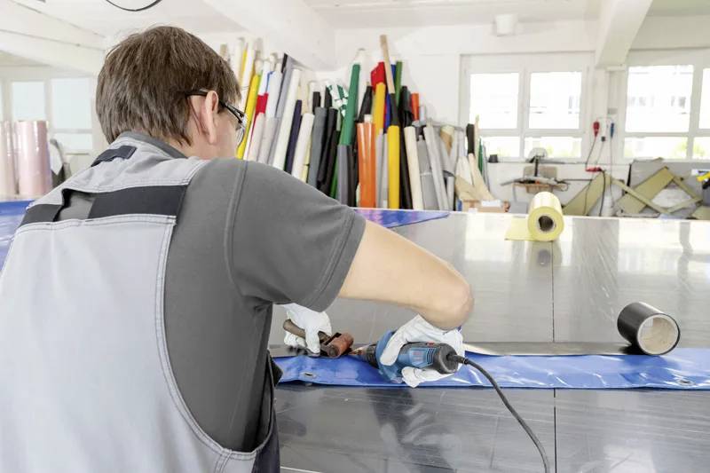 Dělník používá nástroj ke svaření dvou listů modrého materiálu ve výrobní dílně, obklopený rolemi barevných materiálů.