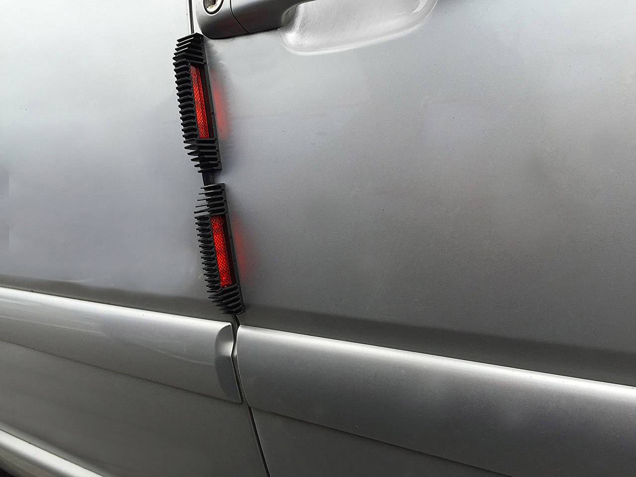 Stříbřitě šedé auto s černými a červenými ochrankami hran dveří na straně řidiče. Lak je lesklý a hladký.