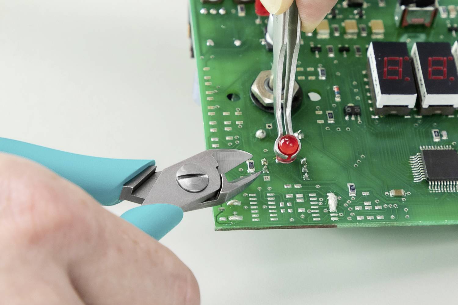Detailní záběr rukou, které manipulují s elektronickou deskou plošných spojů; kleště a pinzeta drží červenou LED diodu v poloze.