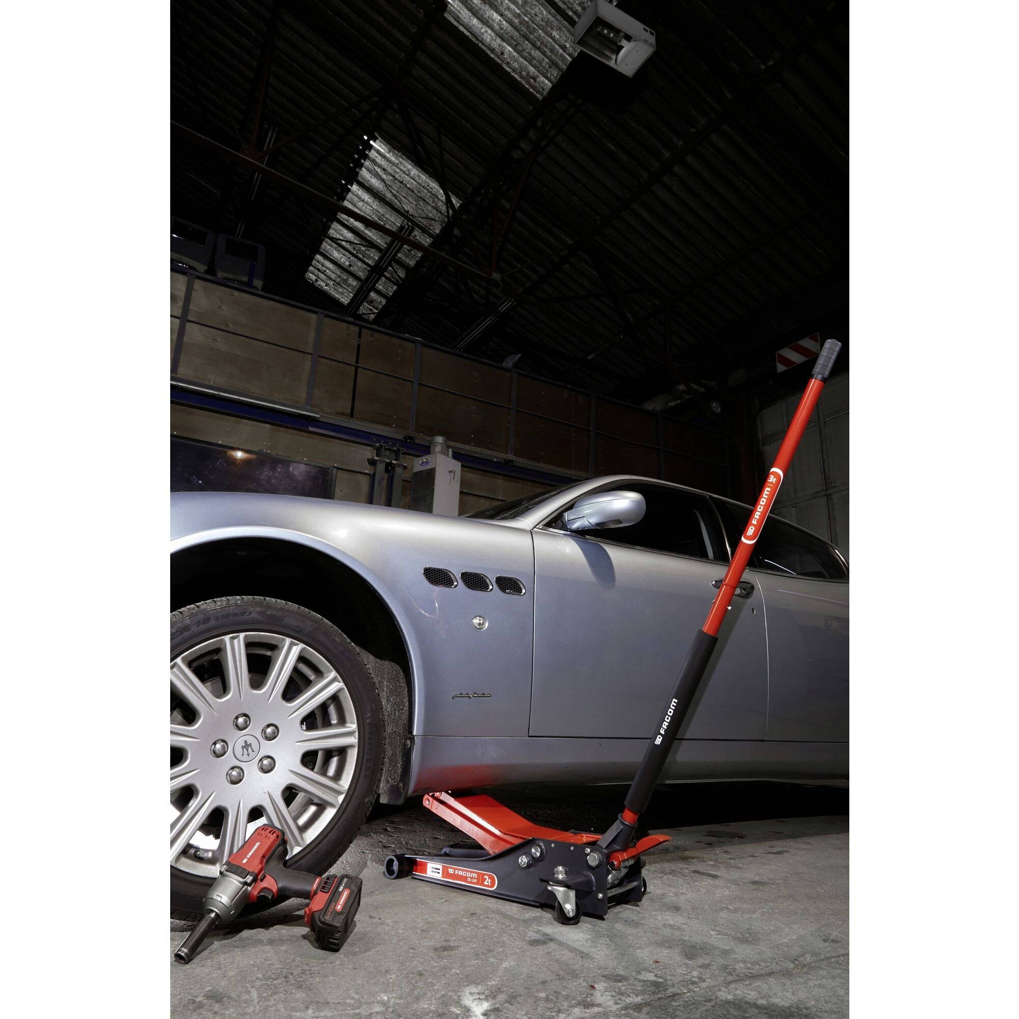 Stříbrné auto je zvedáno hydraulickým zvedákem. V popředí leží nářadí na podlaze garáže.