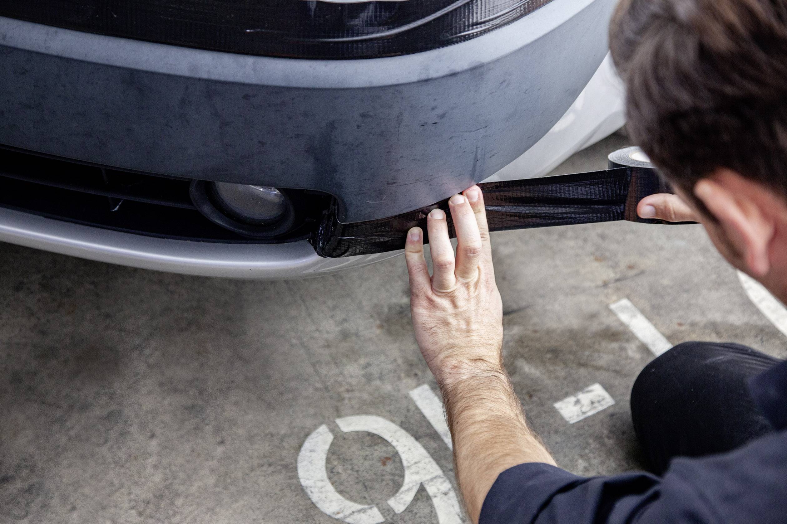 Jedna osoba nalepuje černou fólii na nárazník automobilu v autoservisu, pravděpodobně za účelem opravy nebo vylepšení vzhledu.