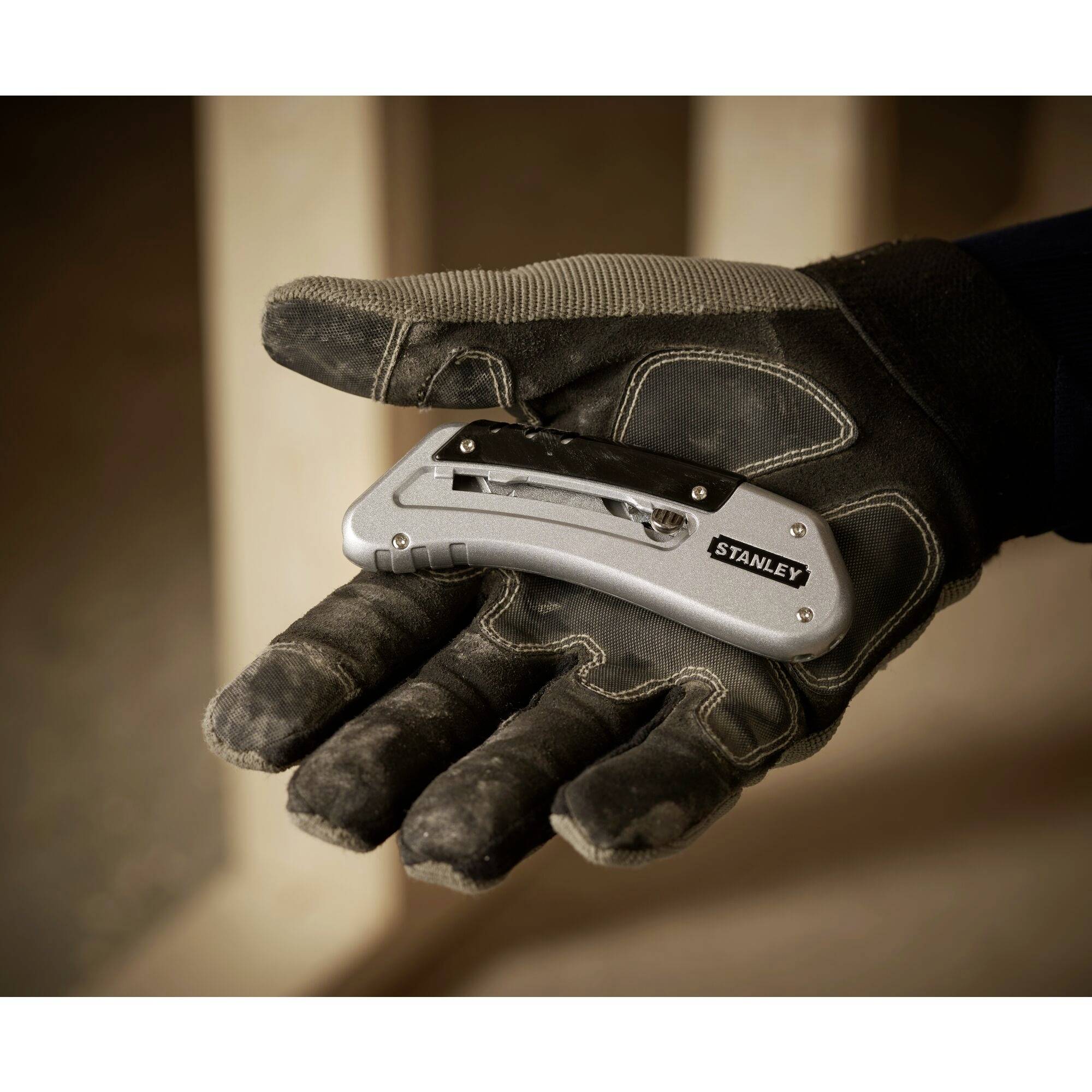 Rukavicí opatěná ruka drží robustní kapesní nůž s nápisem 'Stanley' před rozmazaným pozadím.