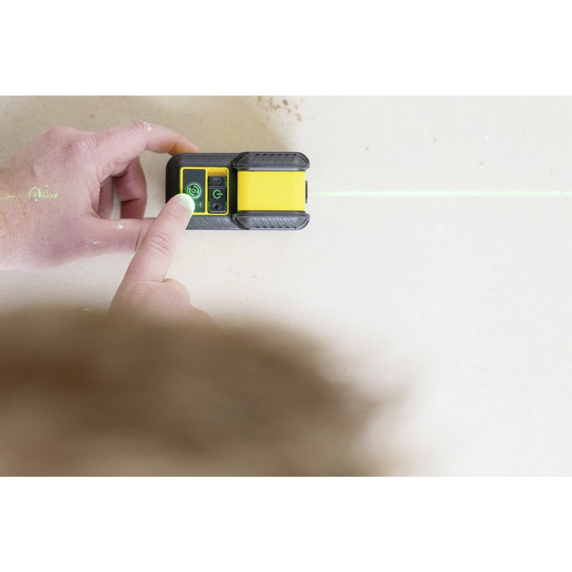 Osoba používá zeďový detektor na světlé stěně. Zelený laserový paprsek vyznačuje přímou linii.