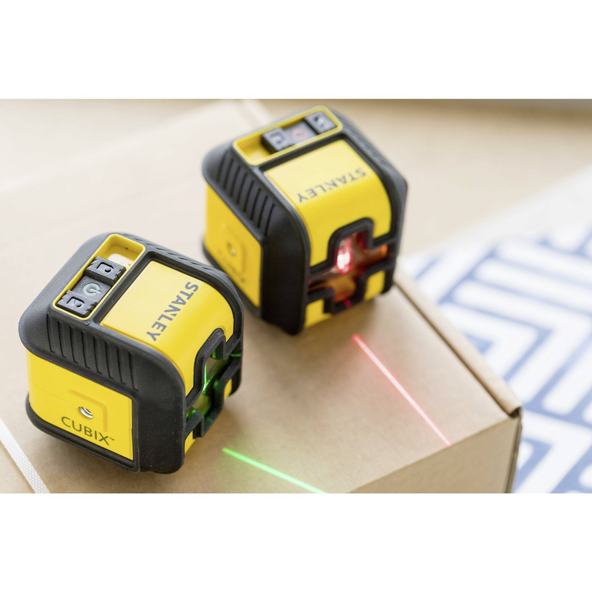 Dva žluté laserové vodováhy promítají vertikální a horizontální zelené, resp. červené laserové čáry. Nacházejí se na kartonu.