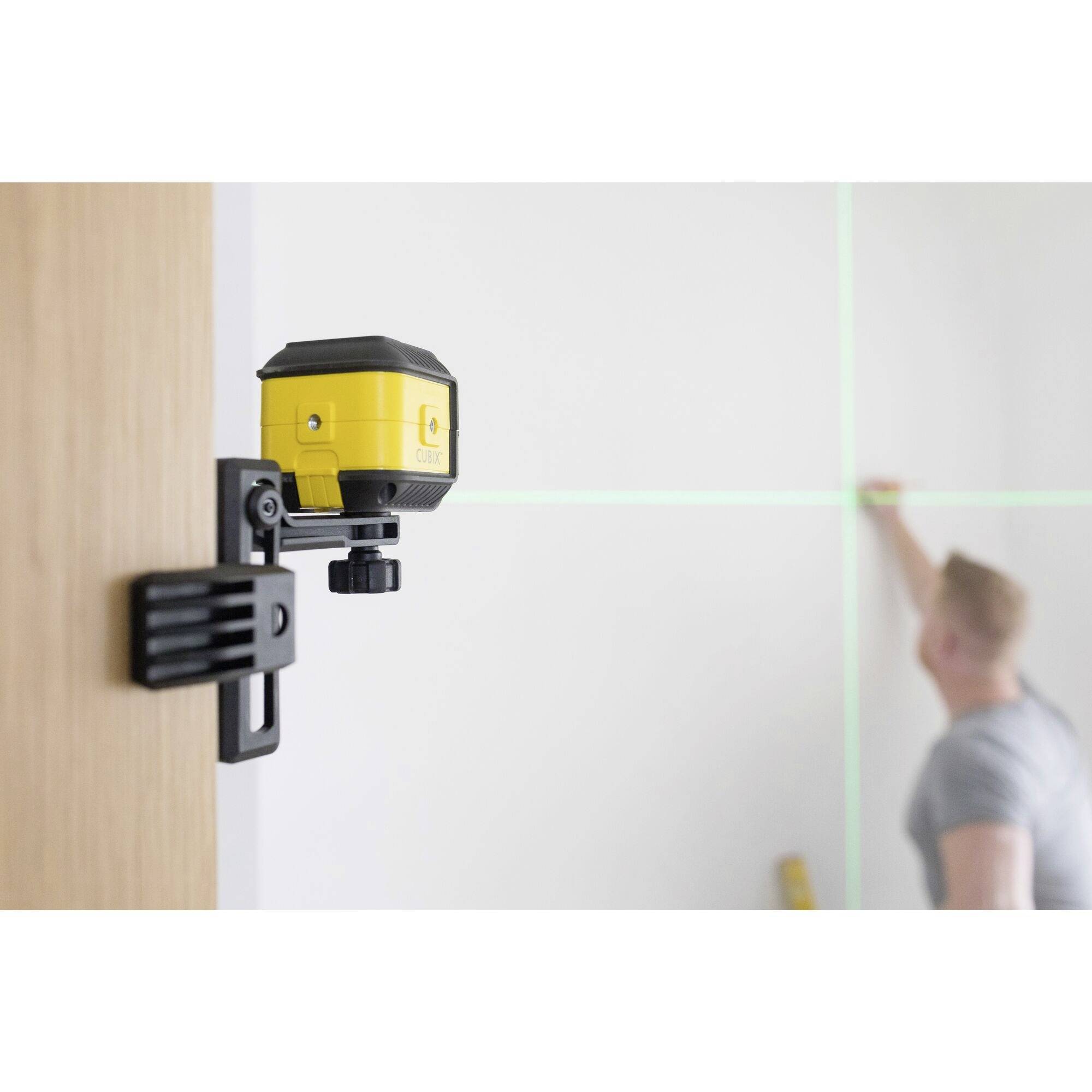 Žlutý křížový čárový laser je namontován na dřevěných dveřích. V pozadí osoba vyznačuje čáru na stěně podle těchto linií.