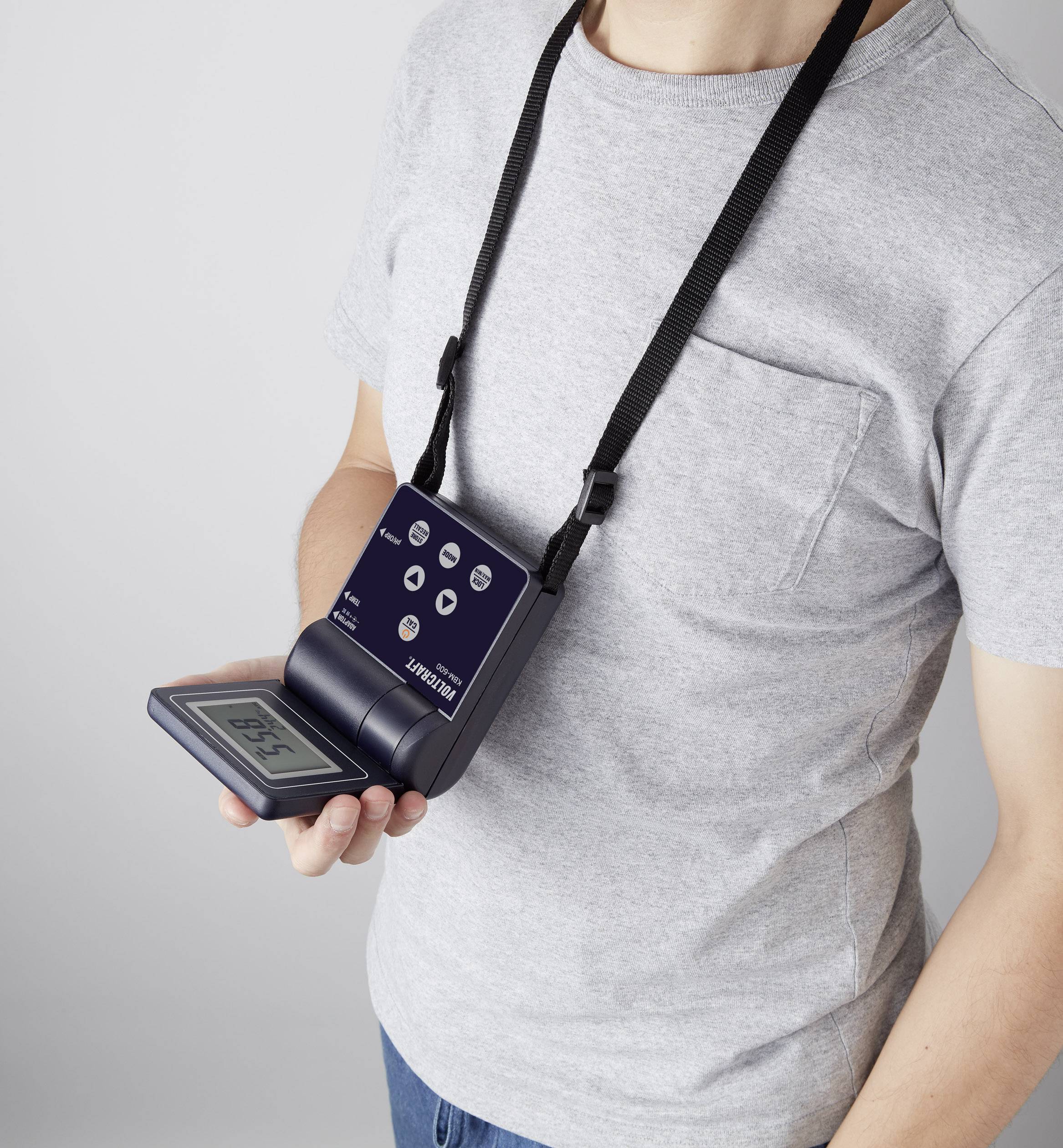 Osoba v šedém tričku drží přenosné elektronické zařízení s digitálním displejem, které je upevněné na šňůrce kolem krku.