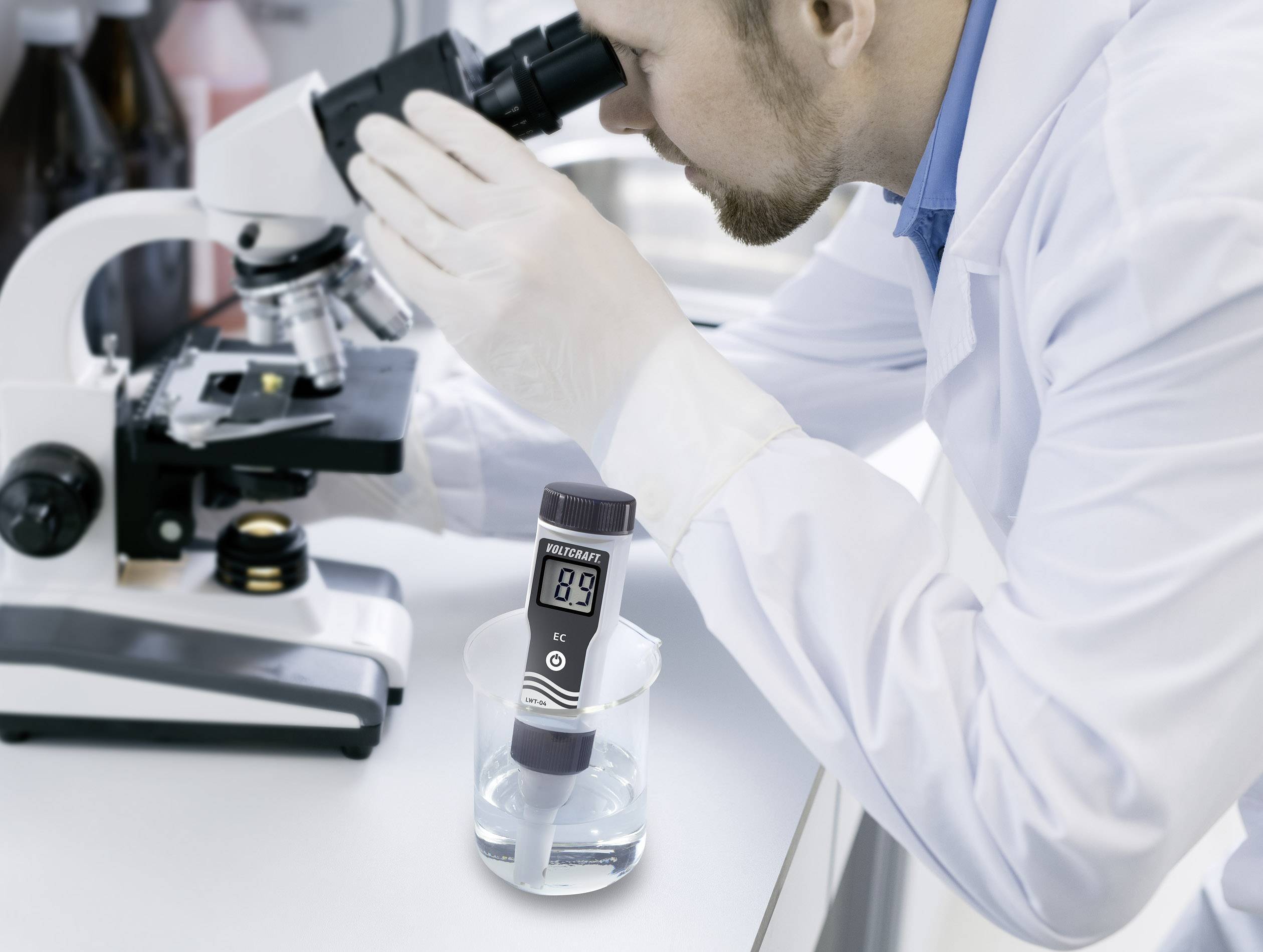 Vědec v laboratorním plášti pozoruje vzorky pod mikroskopem, vedle něj stojí digitální měřicí přístroj v nádobce s kapalinou.