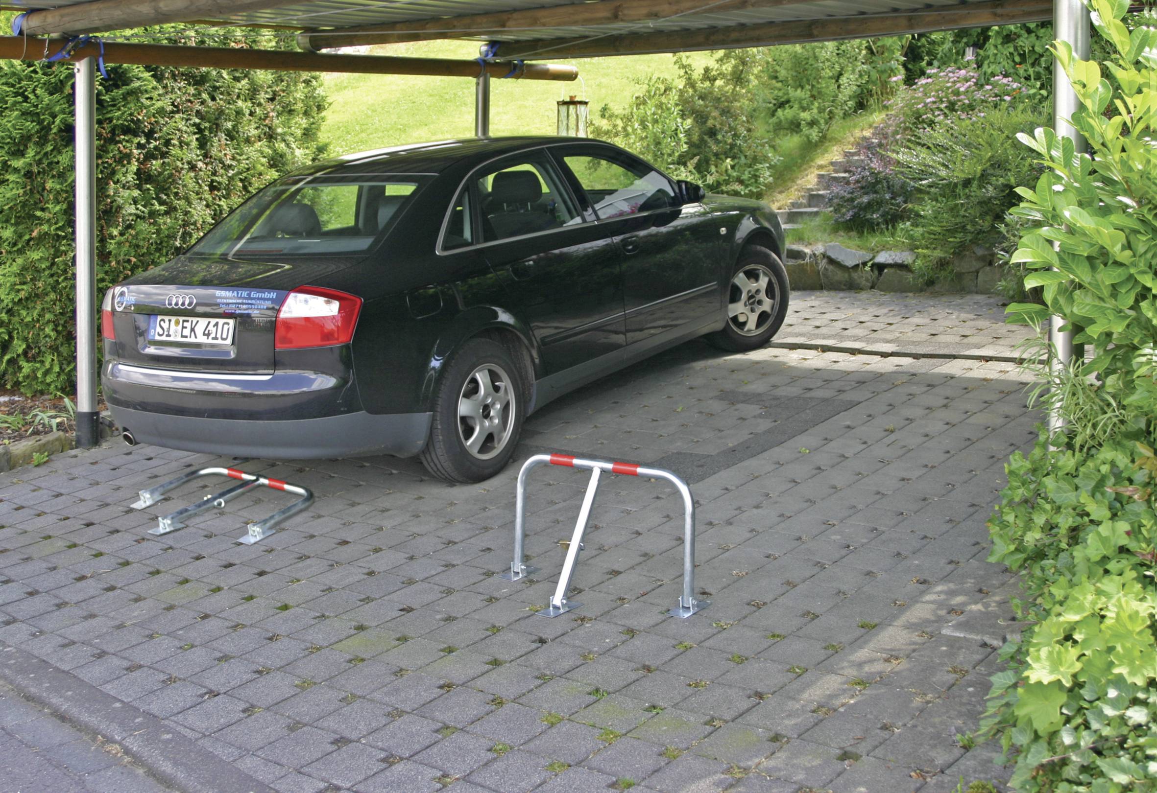 Černé auto je zaparkované pod přístřeškem na dlážděném povrchu. Před vozem jsou vztyčeny dva parkovací sloupky.