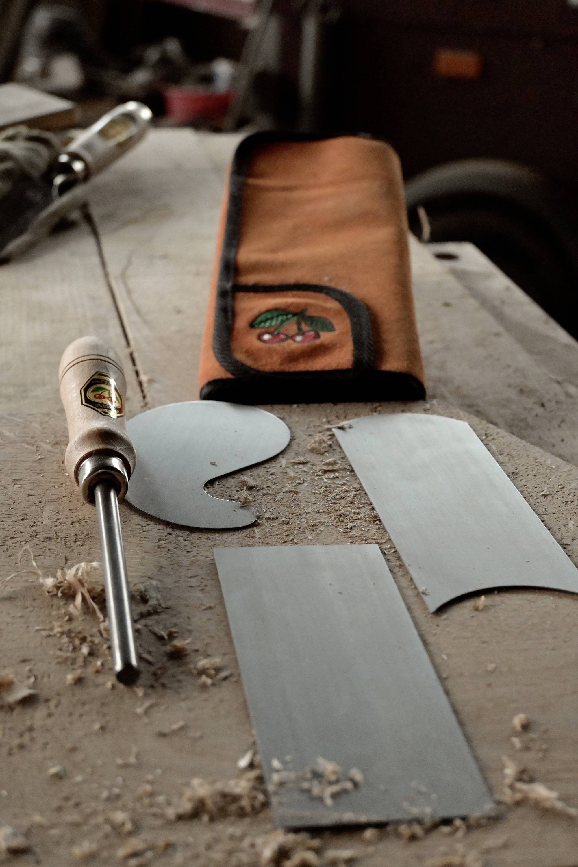 Nástroje na opracování dřeva na stole, včetně ruční škrabky, dvou kovových škrabek, dřevěných pilin a oranžové brašny na nářadí s logem značky Kirsch.