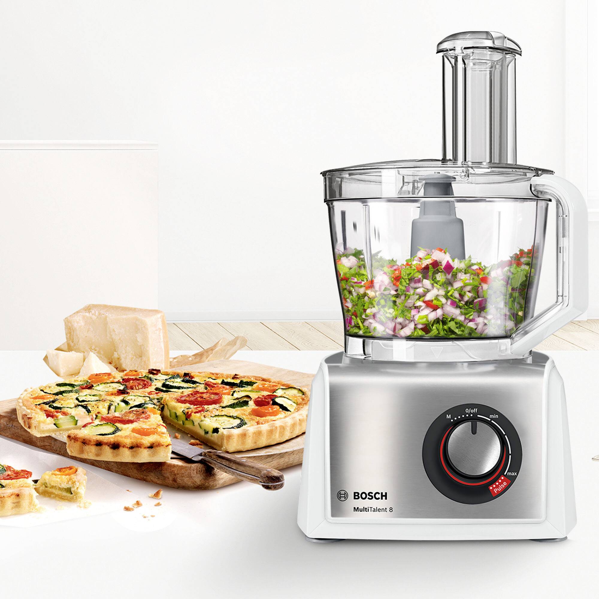 Na dřevěném stole stojí kuchyňský robot s nakrájenou zeleninou v nádobě vedle dvou zeleninových quiche.