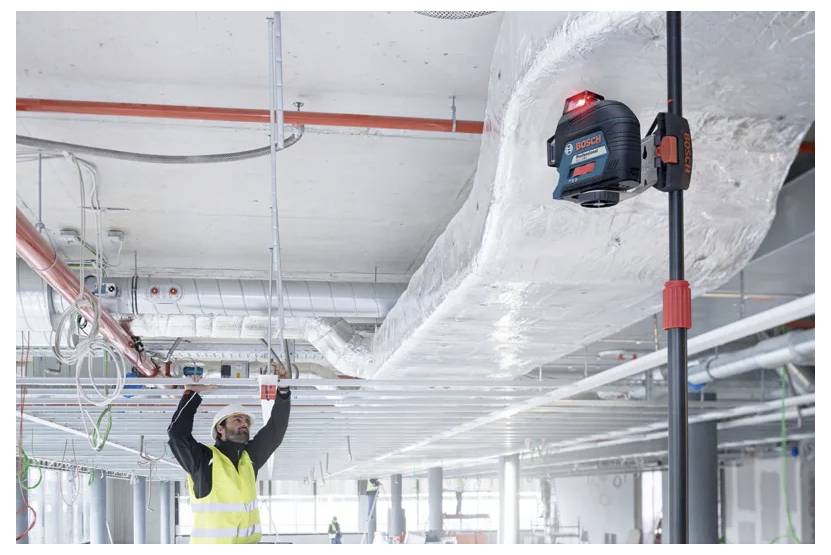 Stavební dělník ve žluté vestě používá laserový nivelační přístroj na staveništi a upravuje stropní instalace ve velké průmyslové budově.