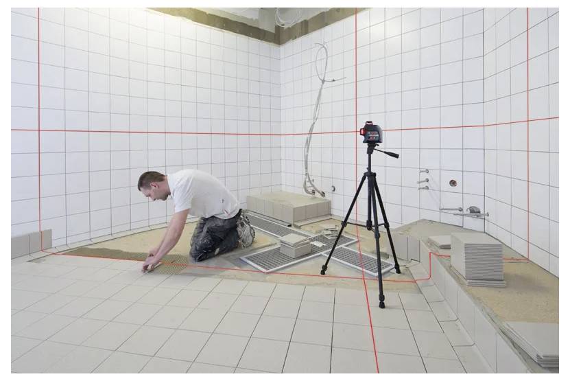 Muž instalující podlahové dlaždice v dlážděné místnosti za použití laserové vodováhy na stativu pro přesné vyrovnání, s nástroji a dlaždicemi viditelnými v okolí.