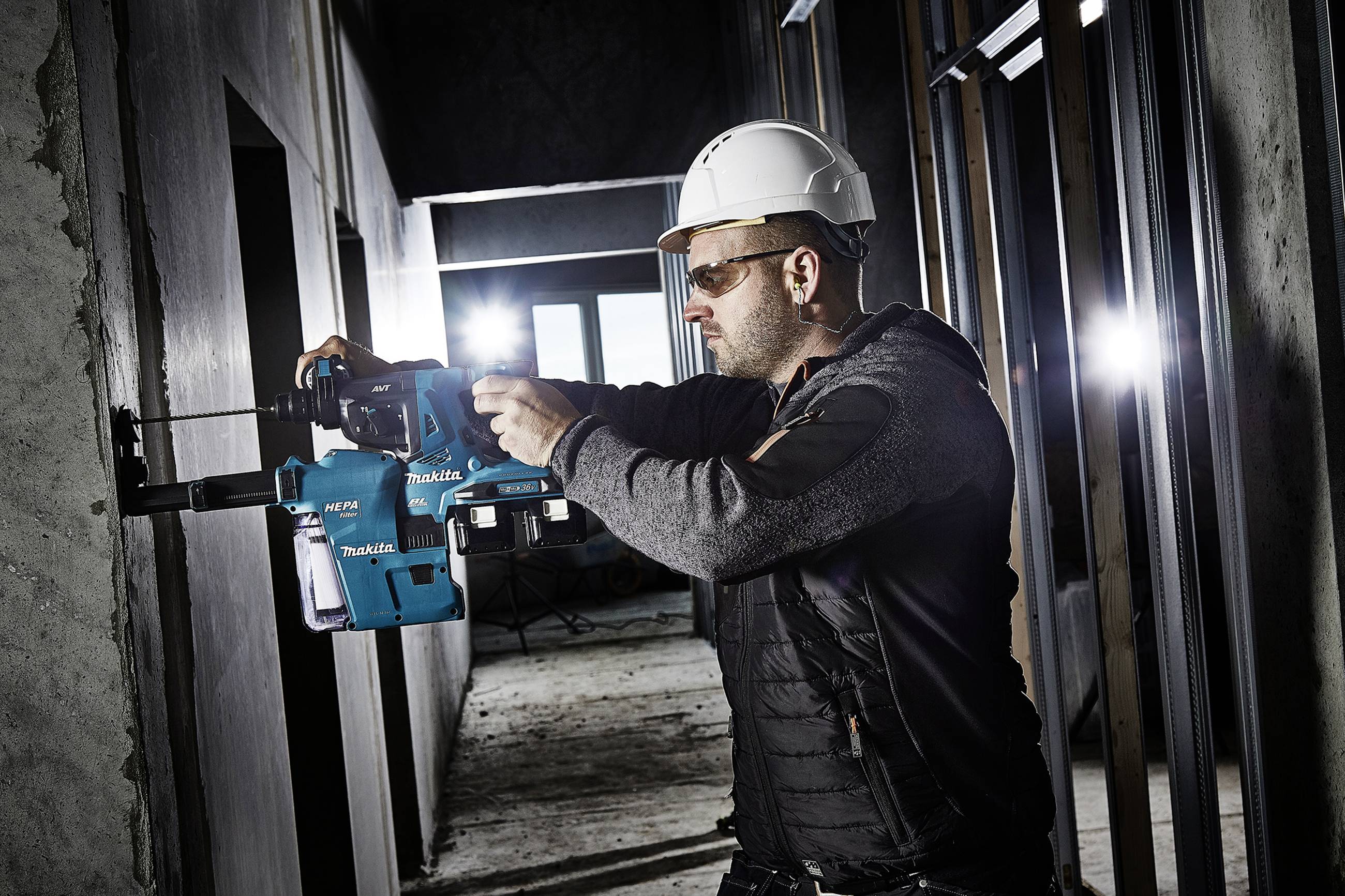 Dělník v ochranné helmě a ochranných brýlích vrtá elektrickým nástrojem do betonové zdi v nedokončené budově.