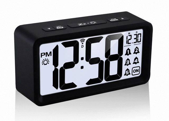 Digitální budík ukazuje čas 12:58 odpoledne. Vedle času jsou na displeji symboly pro alarm, datum a teplotu.