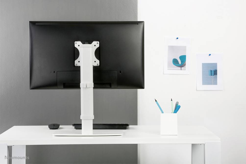 Psací stůl s monitorem na bílém stole. Vedle je nádoba s modrými propiskami. Na stěně visí dva obrazy.