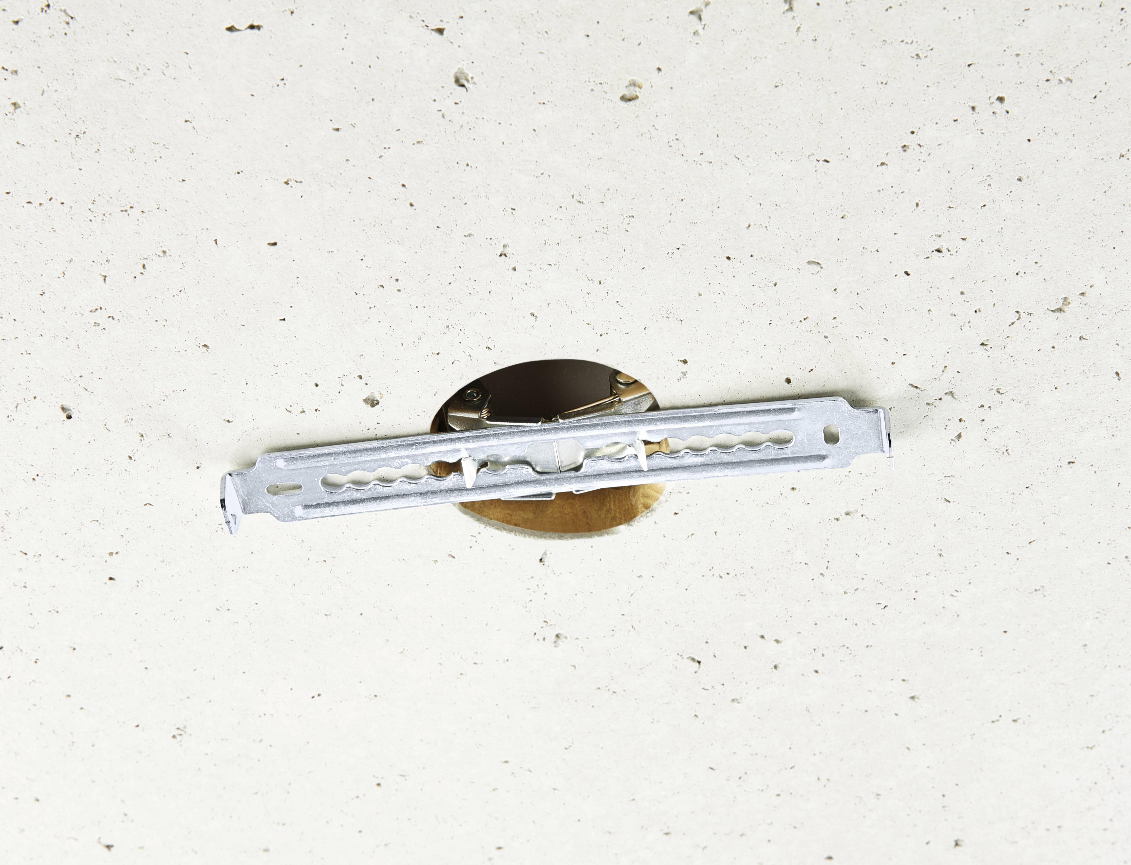 Detailní záběr kovového prvku zabudovaného do bílého povrchu stropu. Vypadá to, že je součástí držáku nebo upevnění.