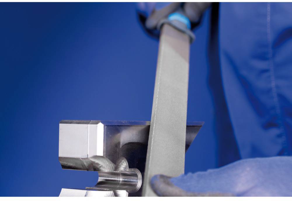 Osoba pilníkem opracovává kovový díl ve svěráku. Má na sobě modré pracovní rukavice a pracovní oděv.