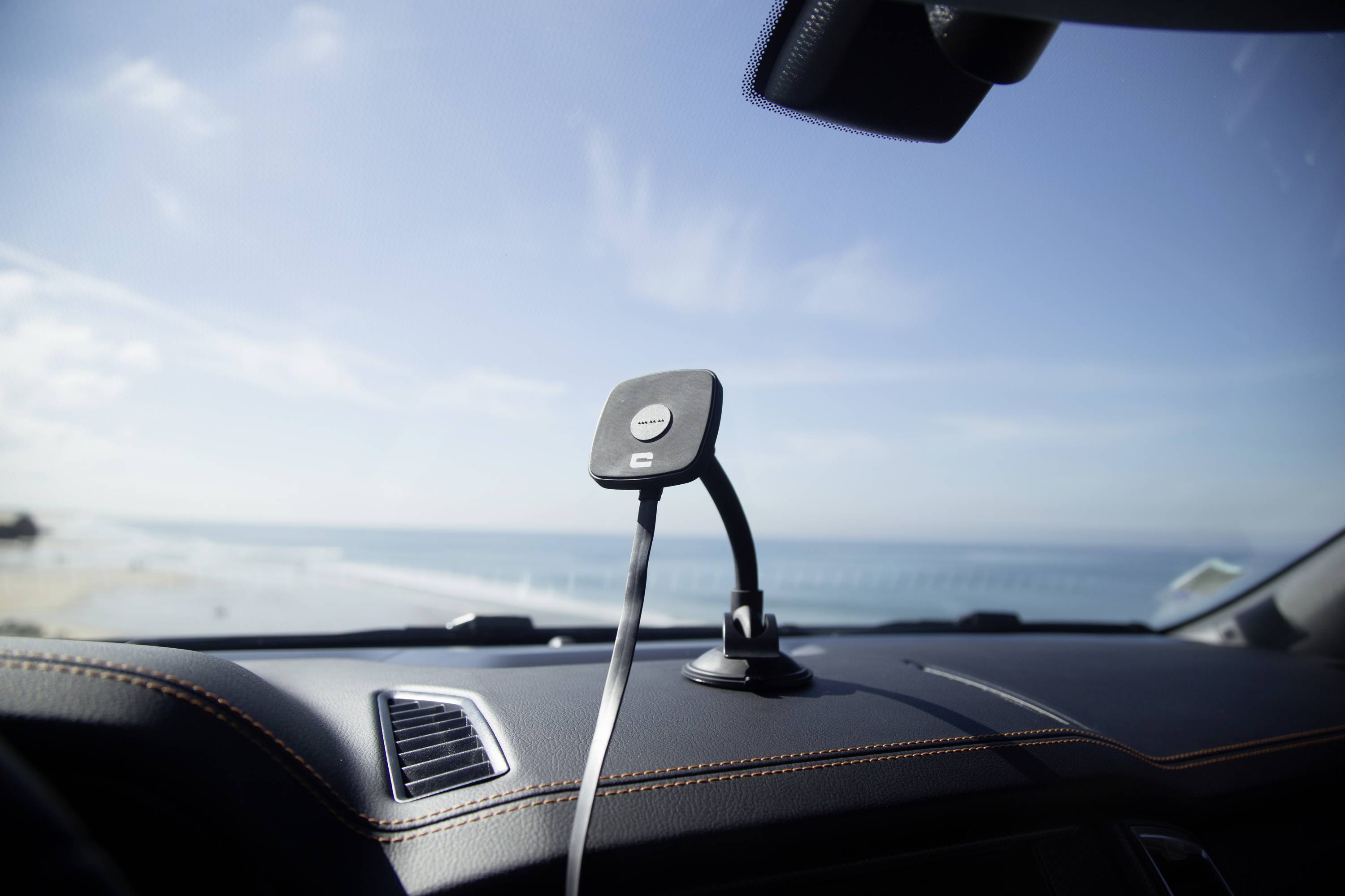 Držák na smartphone je upevněn na palubní desce automobilu. V pozadí je vidět pláž s modrým nebem.