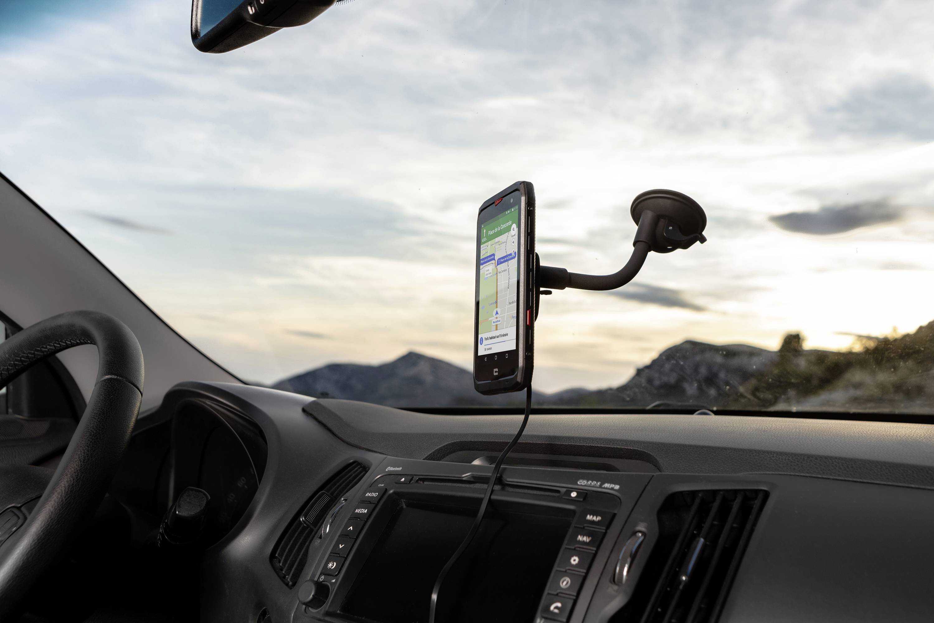 Smartphone na palubní desce zobrazuje navigační aplikaci, přičemž v pozadí jsou vidět hory a obloha s mraky.