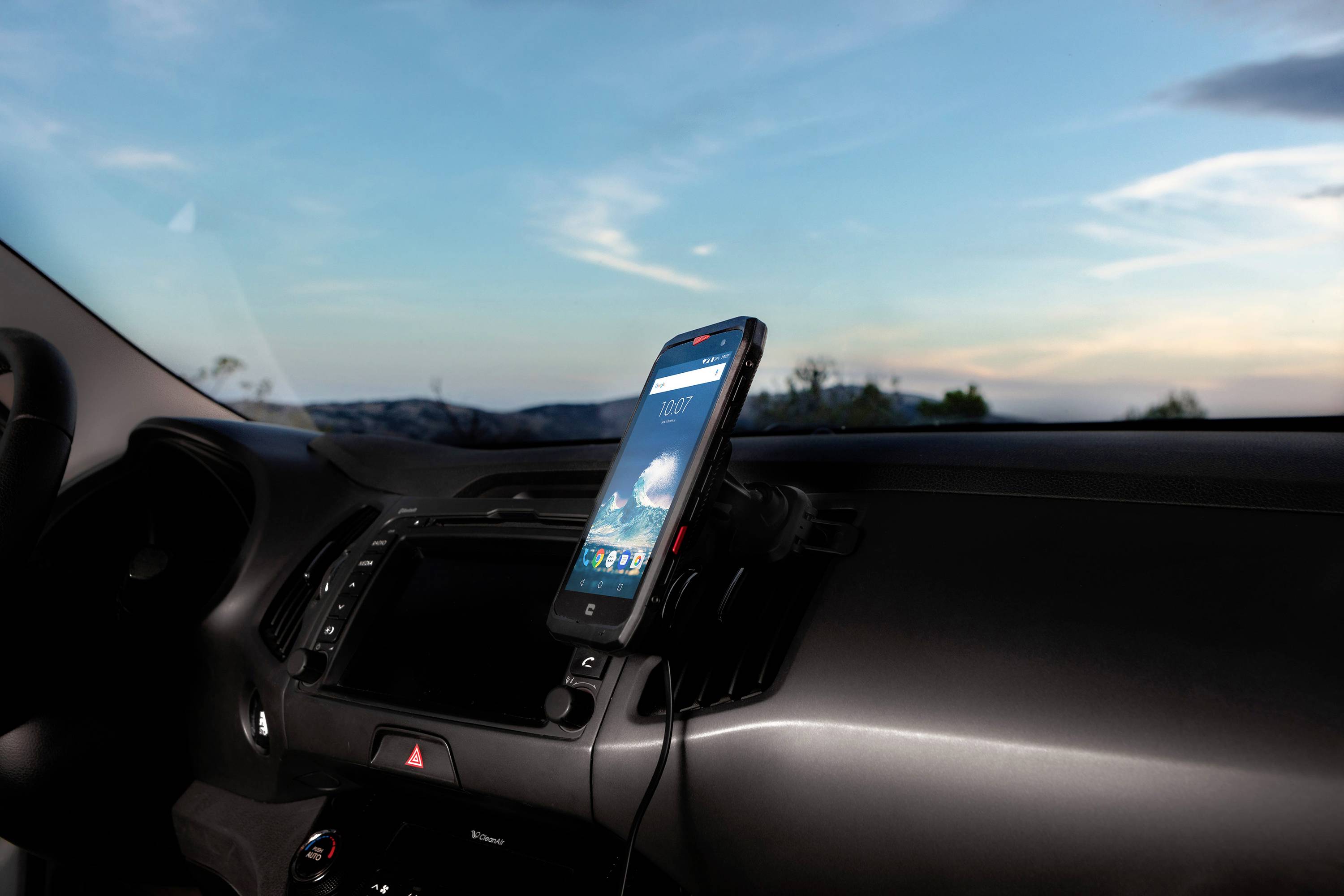 Smartphone v držáku v autě, zobrazující navigační aplikaci, obklopené oblačnou oblohou při západu slunce pohledem skrz čelní sklo.