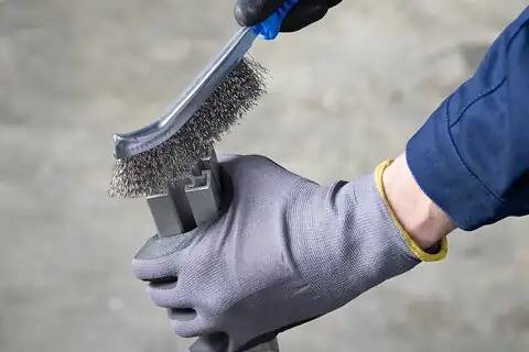 Osoba v rukavicích používá drátěný kartáč k čištění kovové součástky, přičemž se zaměřuje na odstranění rezaviny nebo nečistot, což podtrhuje údržbářské práce.