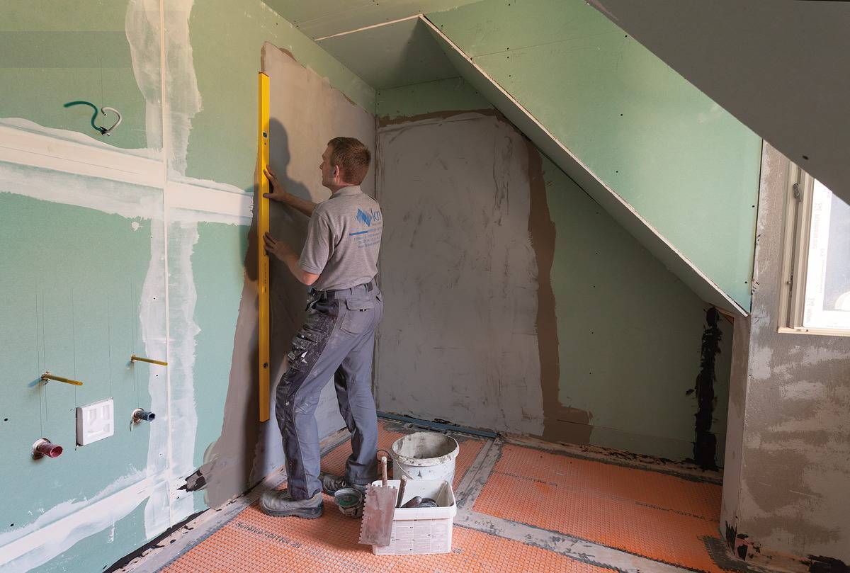 Řemeslník vyrovnává sádrokartonovou stěnu pomocí vodováhy v podkrovním prostoru. Na podlaze jsou rozloženy pracovní materiály.