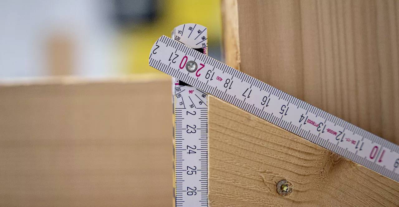 Svinovací metr ukazuje 27 cm u dřevěného kusu v pravém úhlu. Slouží k měření a konstruování.