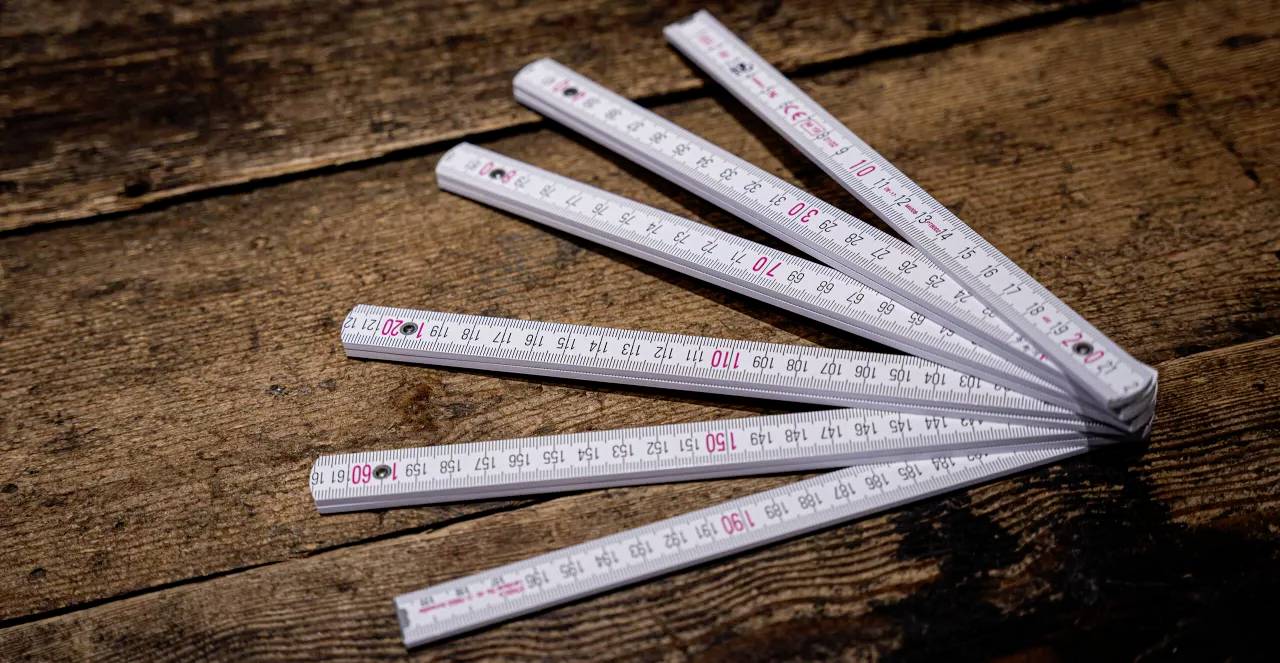 Bílý skládací metr ležící rozložený na dřevěném stole s červenými a černými značkami.