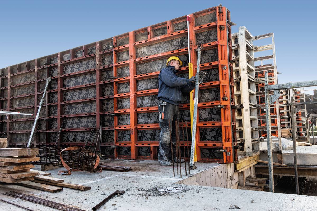 Dělník v žluté přilbě upravuje bednění na staveništi. Stojí na betonovém povrchu obklopen stavebními materiály.
