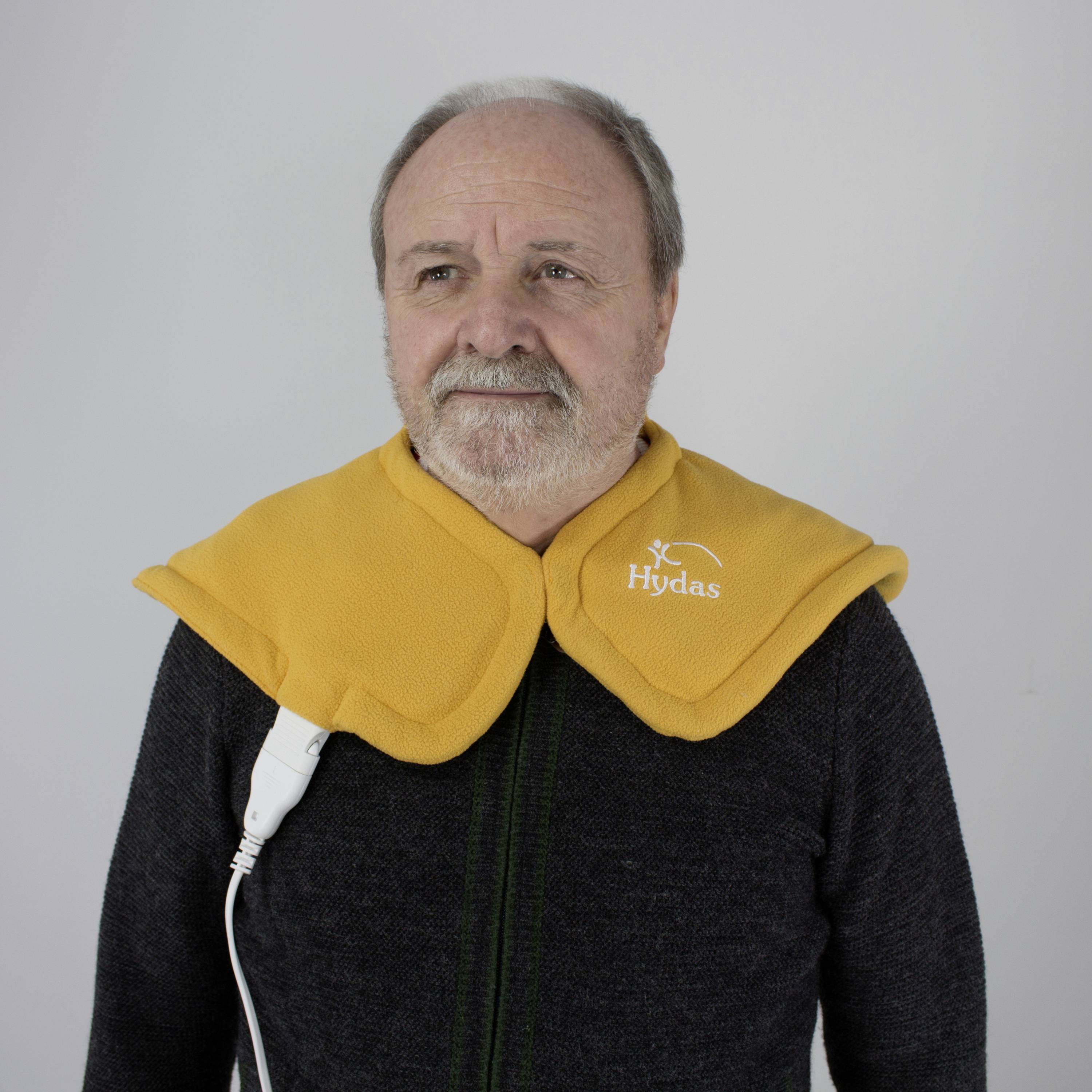 Starší muž nosí vyhřívací krční a ramenní polštářek z žluté látky s integrovaným ovládacím prvkem. Je viditelné firemní logo „Hydas".