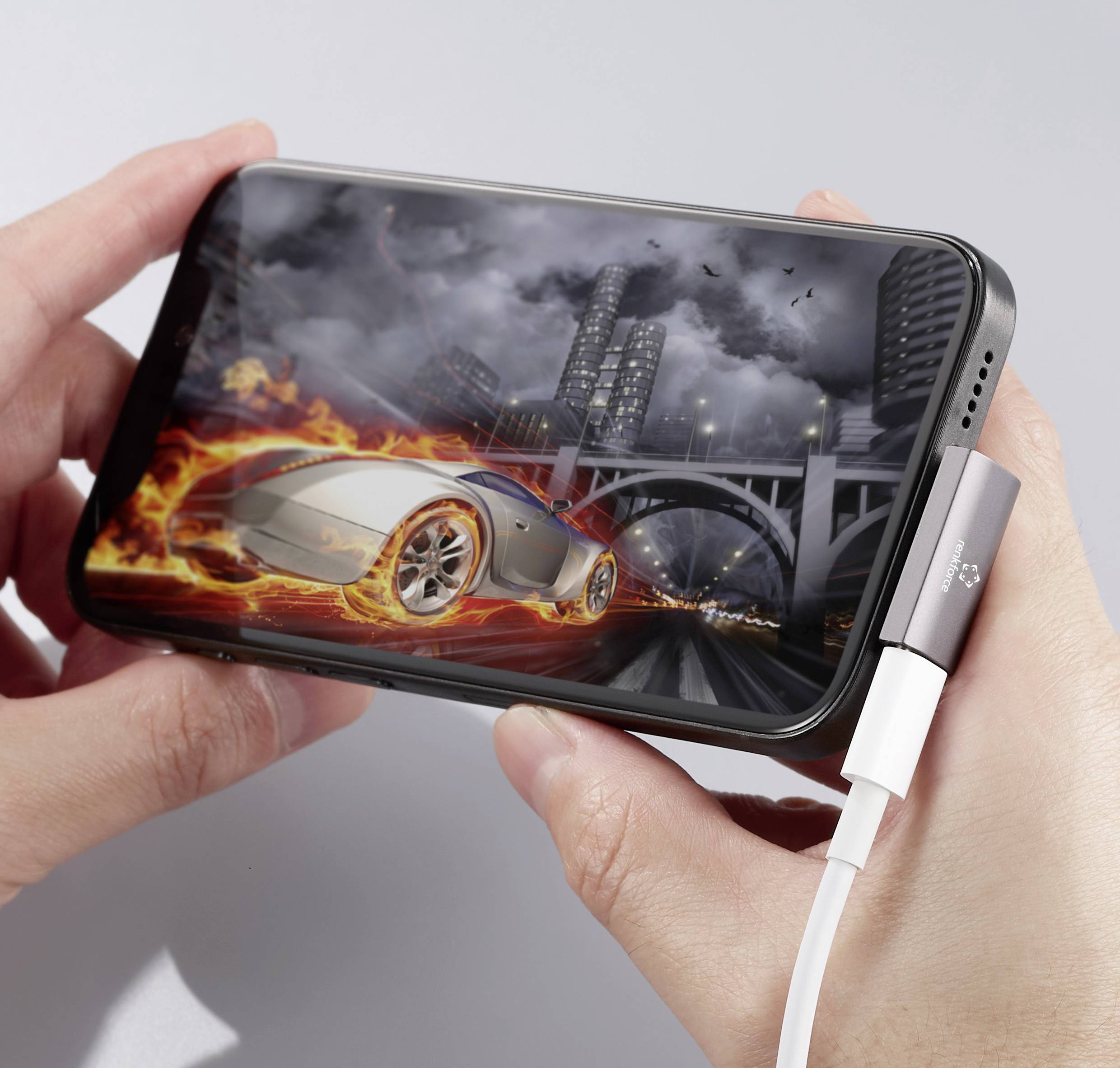Člověk drží smartphone s automobilovou závodní hrou, zobrazuje hořící automobil na městské ulici, zatímco je připojen k nabíjecímu zařízení.