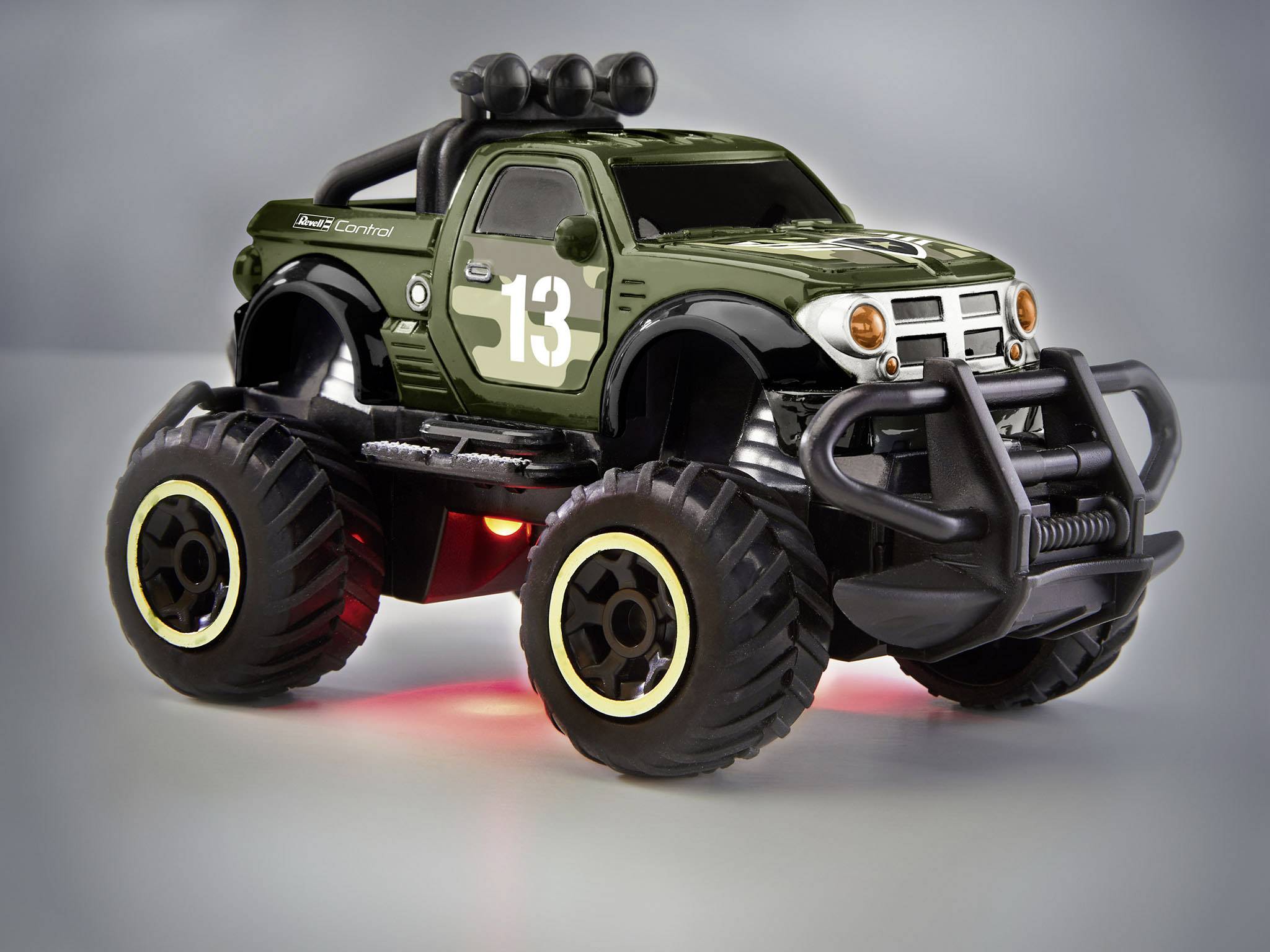 Hračkářský monster truck s číslem 13, olivově zelený, velké pneumatiky, svítící světla, před šedým pozadím.