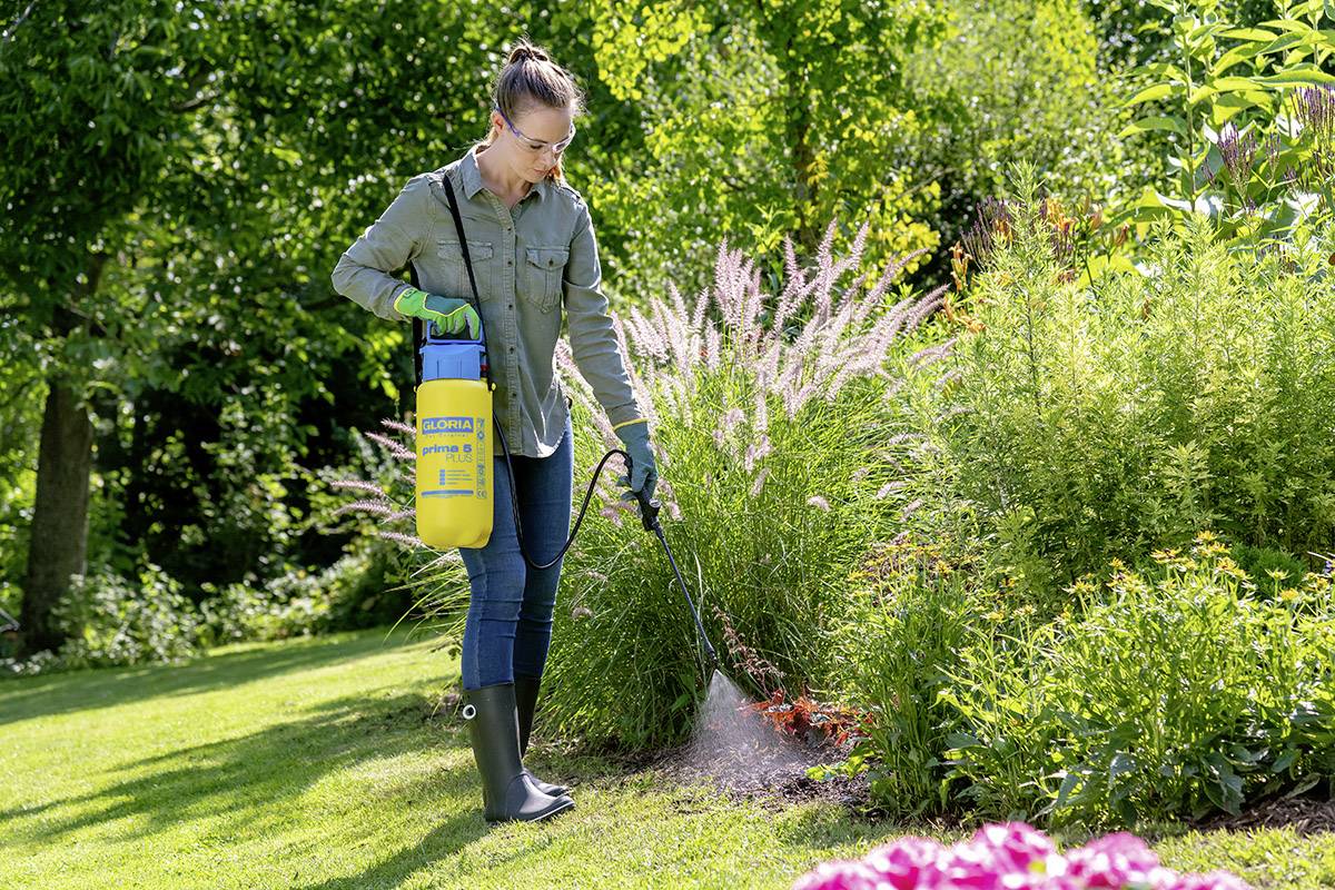Osoba postřikuje pesticidy z žlutého kontejneru na rostliny v slunečné zahradě obklopené zelení.