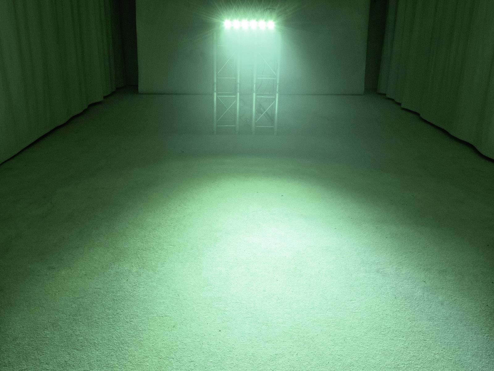 Jasný zelený reflektor osvětluje prázdnou místnost vyloženou kobercem. V pozadí visí závěsy.