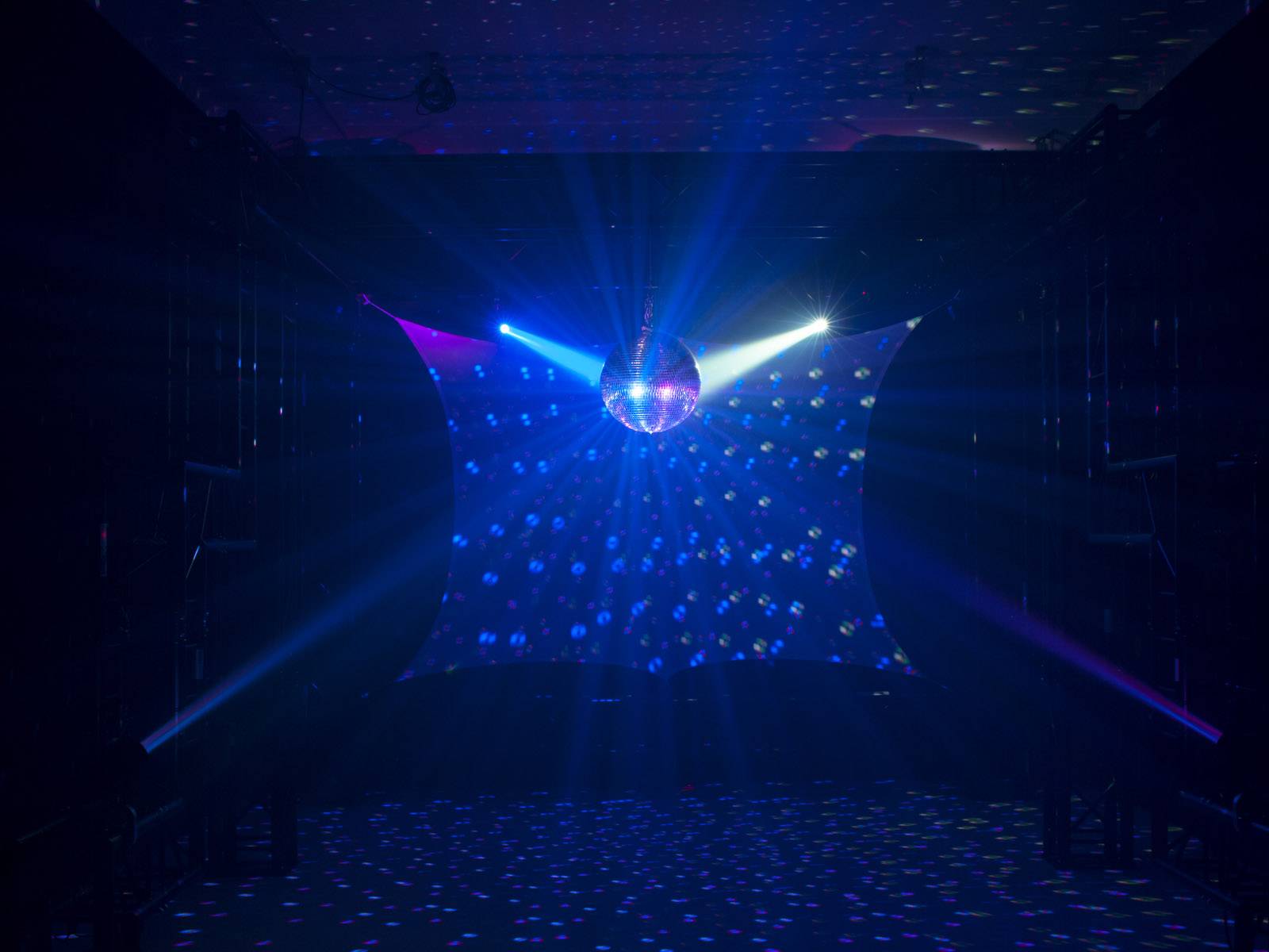 Disco koule v tmavé místnosti, osvětlená barevnými reflektory, vrhá vzory světelných bodů na podlahu a stěny.